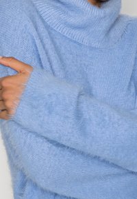 Ljusblå polotröja i mjukt, fluffigt material; har en ribbad krage och ett löst stickat kropp med långa, plyschiga ärmar.