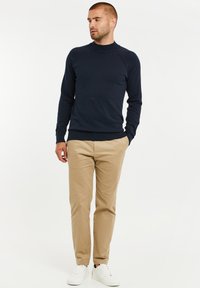 Dunkelblauer Pullover mit hohem Kragen und langen Ärmeln, kombiniert mit hellbeigen Chinos und weißen Sneakers. Glatte Textur, enganliegendes Design.