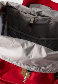 Εσωτερικό κόκκινου σακιδίου πλάτης, με απαλό γκρι ύφασμα, τσέπες με φερμουάρ, ελαστική αποθήκευση και καφέ ετικέτα λογότυπου. Ορατό υφασματισμένο υλικό.