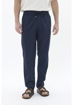 Uomo che indossa pantaloni navy con coulisse e sandali marroni, in piedi contro uno sfondo bianco uniforme.