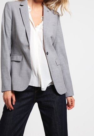 Kvinde iført lysegrå blazer over hvid bluse kombineret med mørkeblå jeans, stående foran en ensfarvet hvid baggrund.