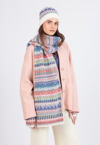 Donna che indossa un cappotto rosa pastello, una sciarpa e un cappello con motivi multicolori, guanti senza dita e pantaloni color crema, che posa su uno sfondo bianco.