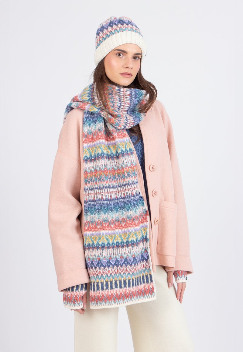 Donna che indossa un cappotto rosa pastello, una sciarpa e un cappello con motivi multicolori, guanti senza dita e pantaloni color crema, che posa su uno sfondo bianco.