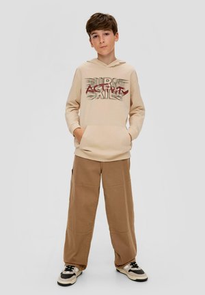 Sudadera beige con un bolsillo frontal y un diseño de texto gráfico. Pantalones cargo sueltos de color marrón claro. Zapatillas negras y blancas con acentos.