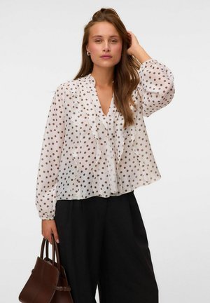 Femme portant un chemisier blanc transparent à pois marron et un pantalon noir, tenant un sac à main marron tout en touchant ses cheveux d'une main.