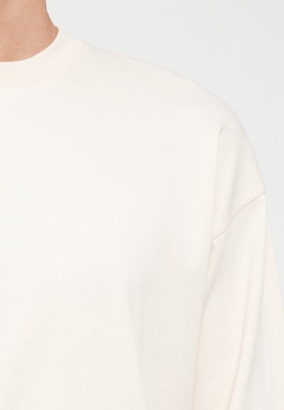 Light beige sweatshirt with a round neckline, dropped shoulders, and smooth cotton texture. Simple design and minimal stitching details.
