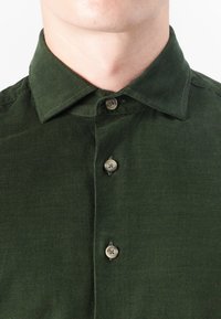 Tumma vihreä corduroy-paita, jossa on terävä kaulus, kahdella nappilla varustettu nappilista ja pystysuora ribbitekstuuri.