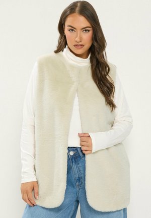 Femme portant une veste sans manches en fausse fourrure couleur crème par-dessus un haut blanc à manches longues et un jean bleu, avec des cheveux bruns ondulés et lâches.