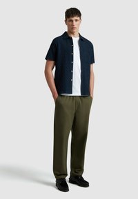 Chemise bleu marine à manches courtes avec un motif texturé, t-shirt blanc en dessous, pantalon vert olive et chaussures noires. Tenue simple et décontractée.
