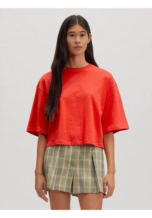 Junge Frau mit langen dunklen Haaren, die ein übergroßes, knallrotes Kurzarmtop und karierte plissierte Shorts trägt, steht vor einem schlichten Hintergrund.