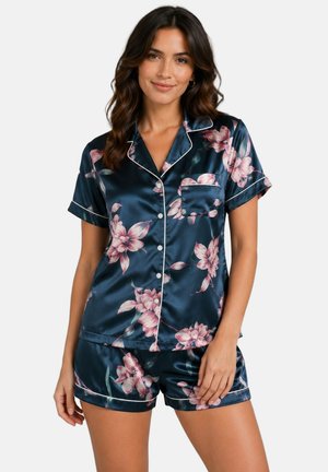 Femme portant un ensemble pyjama en satin bleu marine à manches courtes avec des fleurs roses, un passepoil blanc et un devant boutonné, debout sur fond blanc.
