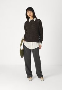 Pull en tricot marron avec un motif texturé, chemise blanche à col en dessous, associé à un pantalon gris et des ballerines beiges. Sac à main vert en accent.
