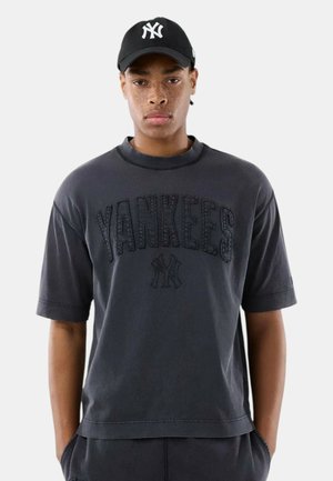 Giovane uomo con lentiggini che indossa un cappellino nero dei Yankees e una T-shirt oversize scura con la scritta "YANKEES" e il logo della squadra sul davanti, mani in tasca.
