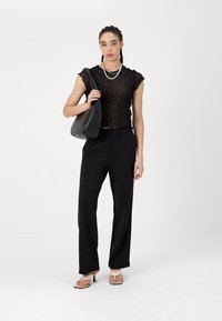 Μαύρο δαντελωτό τοπ με κοντά μανίκια, συνδυασμένο με φαρδιά μαύρα παντελόνια. Το μοντέλο κρατάει μια μαύρη τσάντα ώμου και φοράει μπεζ σανδάλια με τακούνι.