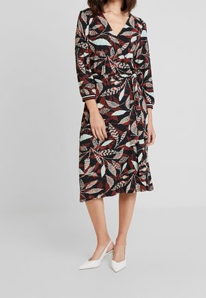 Vrouw die een zwarte wikkeljurk draagt met rood, beige en lichtblauwe bladpatronen, driekwart mouwen en witte pumps met een puntige neus.