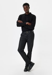 Zwart longsleeve polo shirt gecombineerd met zwarte broek. Model draagt een zwarte riem en sneakers, waardoor een strakke monochrome uitstraling ontstaat.