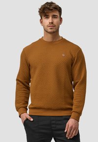 Brauner Sweatshirt mit Textur, rundem Ausschnitt und gerippten Bündchen. Verfügt über ein kleines Logo-Detail auf der Brust. Getragen mit schwarzen Hosen.