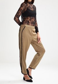 Donna che indossa un top lungo a maniche nere in pizzo trasparente, pantaloni in satin beige con strisce nere ai lati e décolleté neri a punta, in posa di lato.