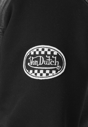 Tissu noir avec un patch ovale blanc présentant un motif en damier et le texte brodé « Von Dutch » en écriture stylisée.