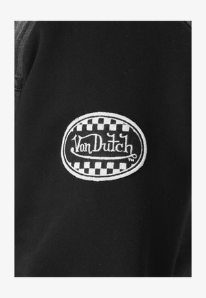 Tissu noir avec un patch ovale blanc présentant un motif en damier et le texte brodé « Von Dutch » en écriture stylisée.