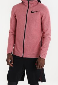 Rote Zip-Hoodie aus einem weichen, strukturierten Stoff mit schwarzen Akzenten, ausgestattet mit einem schwarzen Nike-Logo auf der Brust, kombiniert mit schwarzen Shorts.