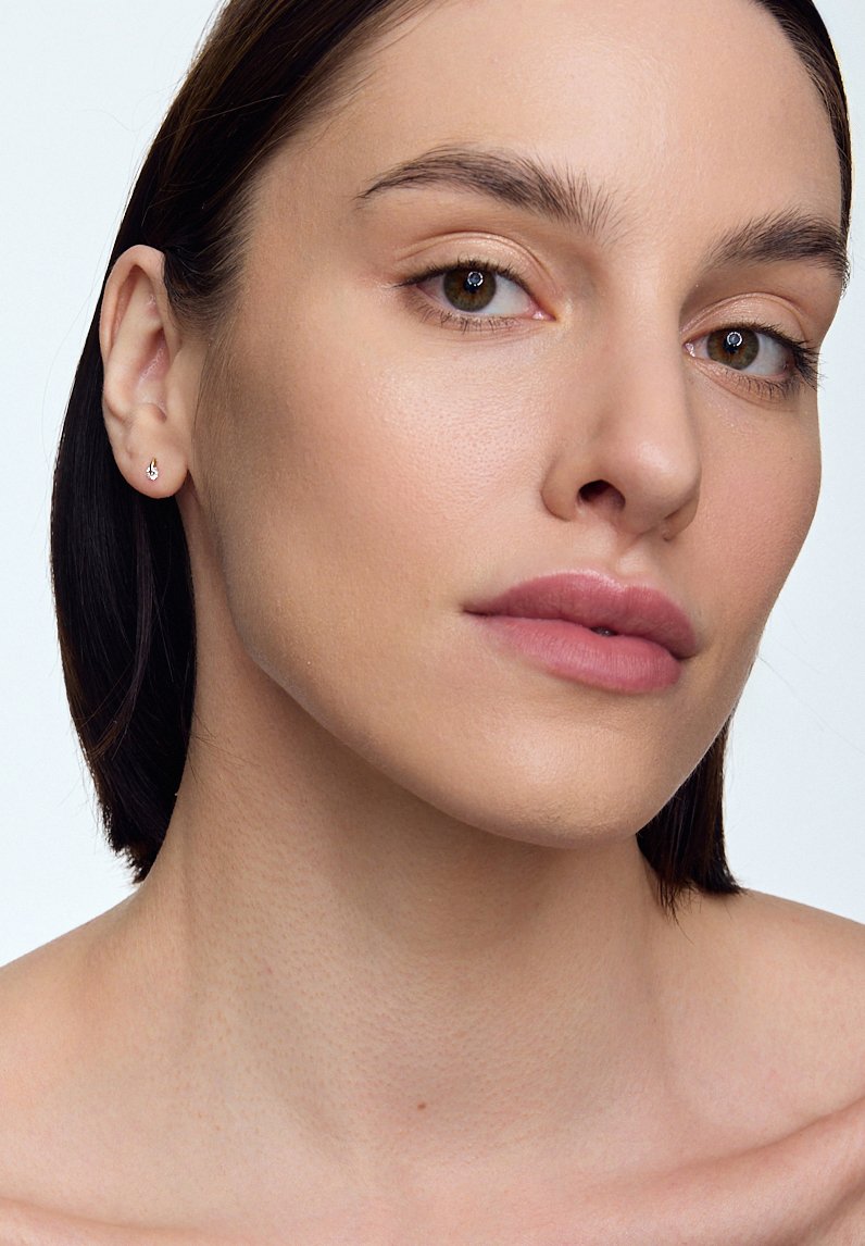 Srebrna kolczyk w przekutym uchu, gładka skóra, neutralny kolor ust, minimalny makijaż i włosy ciemne do ramion na jednolitym tle.