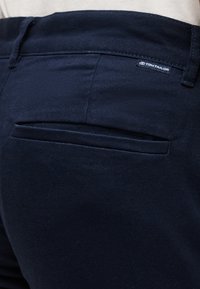 Σκούρο μπλε παντελόνια με μια πίσω τσέπη με κουμπιά και μια μικρή ετικέτα της μάρκας "Tom Tailor" πάνω από την τσέπη.