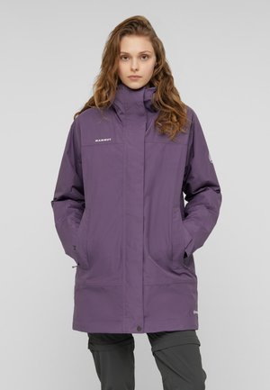 Giovane donna che indossa una lunga giacca viola Mammut con le mani nelle tasche, in piedi davanti a uno sfondo grigio chiaro uniforme.