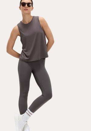 Kvinde iført mørkegrå ærmeløs top, matchende leggings, hvide sokker med striber, hvide sneakers og solbriller, poserende med hænderne bag ryggen.