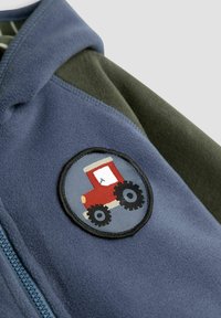 Sinine ja roheliselt fliisjakki ümmargune plaastriga, millel on rautšitud punane traktor suurte mustade ratastega rinnas. 