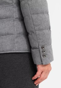 Manga de chaqueta acolchada gris con tres botones; tejido texturizado; un diseño acolchado; dobladillo de manga más oscuro en contraste. Mano reposando sobre una superficie más oscura.