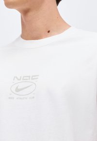 Άτομο που φοράει άσπρο μπλουζάκι Nike Athletic Club με μικρό λογότυπο στο στήθος, δείχνοντας ένα φακί στο λαιμό σε απλό φόντο.