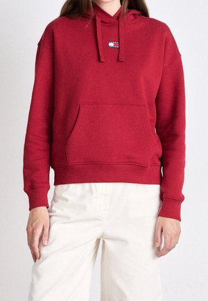 Nainen pukeutuneena punaiseen huppariin, jossa on etutasku ja "Tommy Jeans" -logo, yhdistettynä off-white-värisiin housuihin yksinkertaisella taustalla.