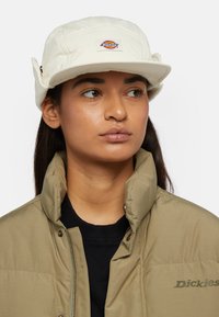 Weißer Bucket Hat mit flachem Rand und gesticktem Logo, kombiniert mit einer khakifarbenen Pufferjacke und einem schwarzen Sweatshirt. Glatte Stofftexturen sind sichtbar.
