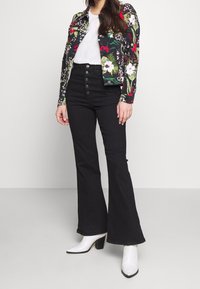 Giacca di jeans con motivo floreale in verde, rosso e bianco; jeans a vita alta aderenti e svasati neri; stivali bianchi con tacco.