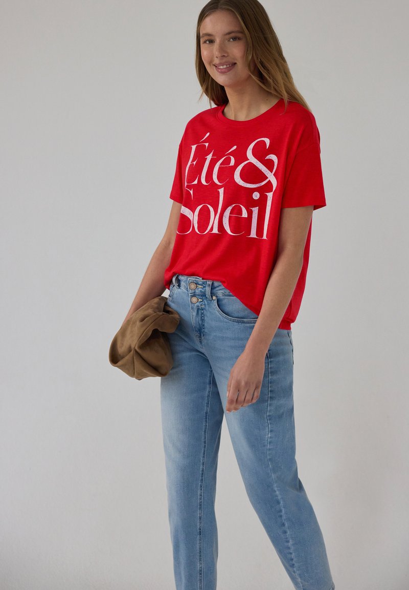 Joven sonriente, vestida con una camiseta roja brillante con el texto "Été & Soleil", vaqueros azul claro, sosteniendo una chaqueta marrón doblada en una mano.