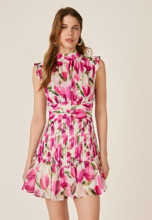 Mujer con vestido rosa y blanco sin mangas con volantes y estampado floral, con cuello alto y falda plisada, de pie frente a un fondo liso.