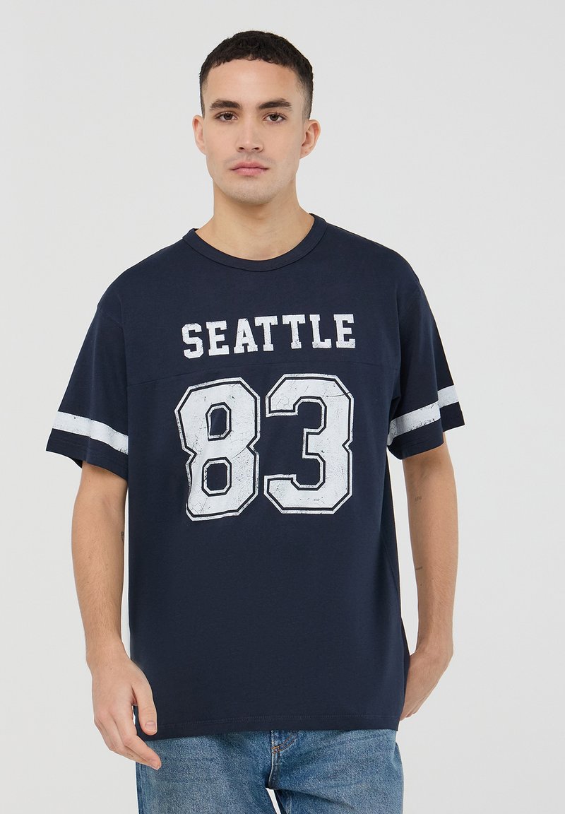 T-shirt in cotone blu navy con maniche corte, caratterizzato dal testo bianco "SEATTLE" e dal numero "83". Stampa testurizzata, vestibilità casual, design minimalista.