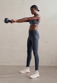 Schwarze Sportleggings mit einem strukturierten Muster, kombiniert mit einem grauen Sport-BH und hellen Sportschuhen, die eine Kettlebell in der rechten Hand halten.