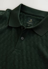 Tamnozelena polo majica s teksturom, dva gumba i rebrasti ovratnik, regular fit, veličina srednja, vidljiva oznaka marke unutar ovratnika.