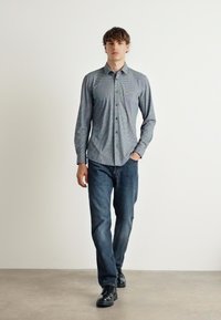 Srajca z gumbom in geometrijskim vzorcem, dolgimi rokavi in ovratnikom; v kombinaciji s temno modrimi jeans hlačami in črnimi čevlji. Enobarvna ozadje.