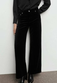 Pantalones de terciopelo negro de pierna ancha con una textura suave, cintura alta, bolsillos frontales y detalles en metal plateado. Combinados con botas negras de tobillo.