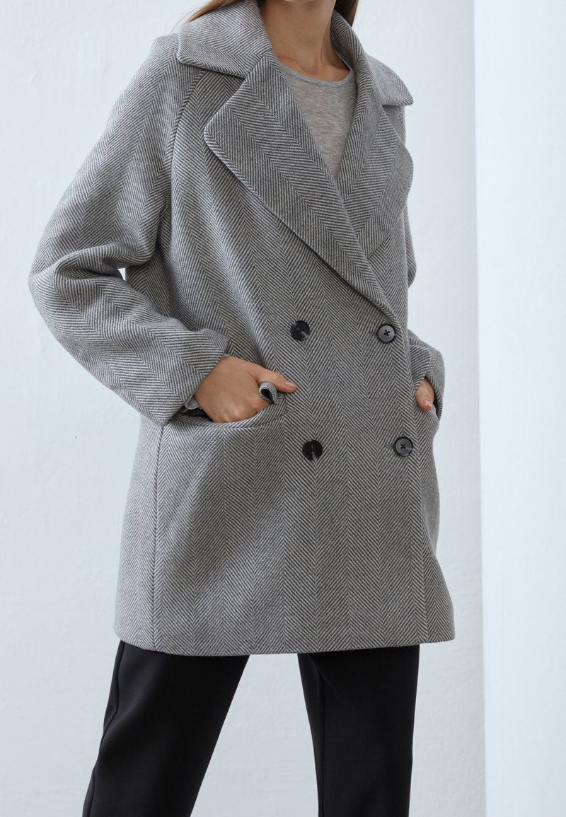 Femme portant un manteau gris à double boutonnage en chevrons avec de grands revers et des boutons noirs, les mains dans les poches, assorti à un pantalon noir.