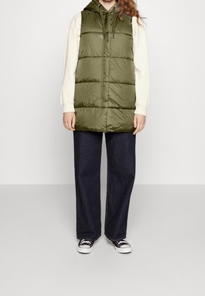 Personne portant une doudoune vert olive avec capuche, un haut crème à manches longues, un jean foncé à jambes larges et des baskets noires et blanches.