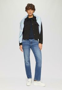 Schwarze kurze Jacke mit Knopfleiste, hellblauer Schal über die Schultern drapiert, marineblaues Oberteil und mittelblaue gerade Jeans, kombiniert mit weißen Sneakern.