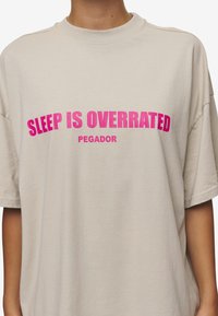 Beige Baumwoll-T-Shirt mit kurzen Ärmeln. Auf der Vorderseite befindet sich auffälliger pinker Text: "SCHLAF IST ÜBERBEBERT" und darunter in kleinerer Schrift "PEGADOR".