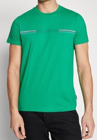 Zielona bawełniana koszulka z okrągłym dekoltem, krótkimi rękawami i poziomym paskiem z logo w kolorze niebieskim i białym na piersi.