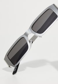 Sonnenbrille mit einem silbernen Metallrahmen und großen schwarzen Gläsern. Enthält klare Nasenpolster und schwarze Bügel mit weißer Schrift.