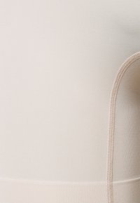 Beige naadloos stoffen kledingstuk met een ribbelstructuur en een gladde afwerking, met contrasterende stiksels langs de randen.