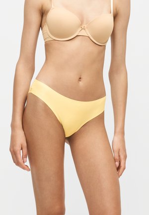 Femme portant un soutien-gorge beige et un slip bikini jaune sans coutures sur un fond clair uni.